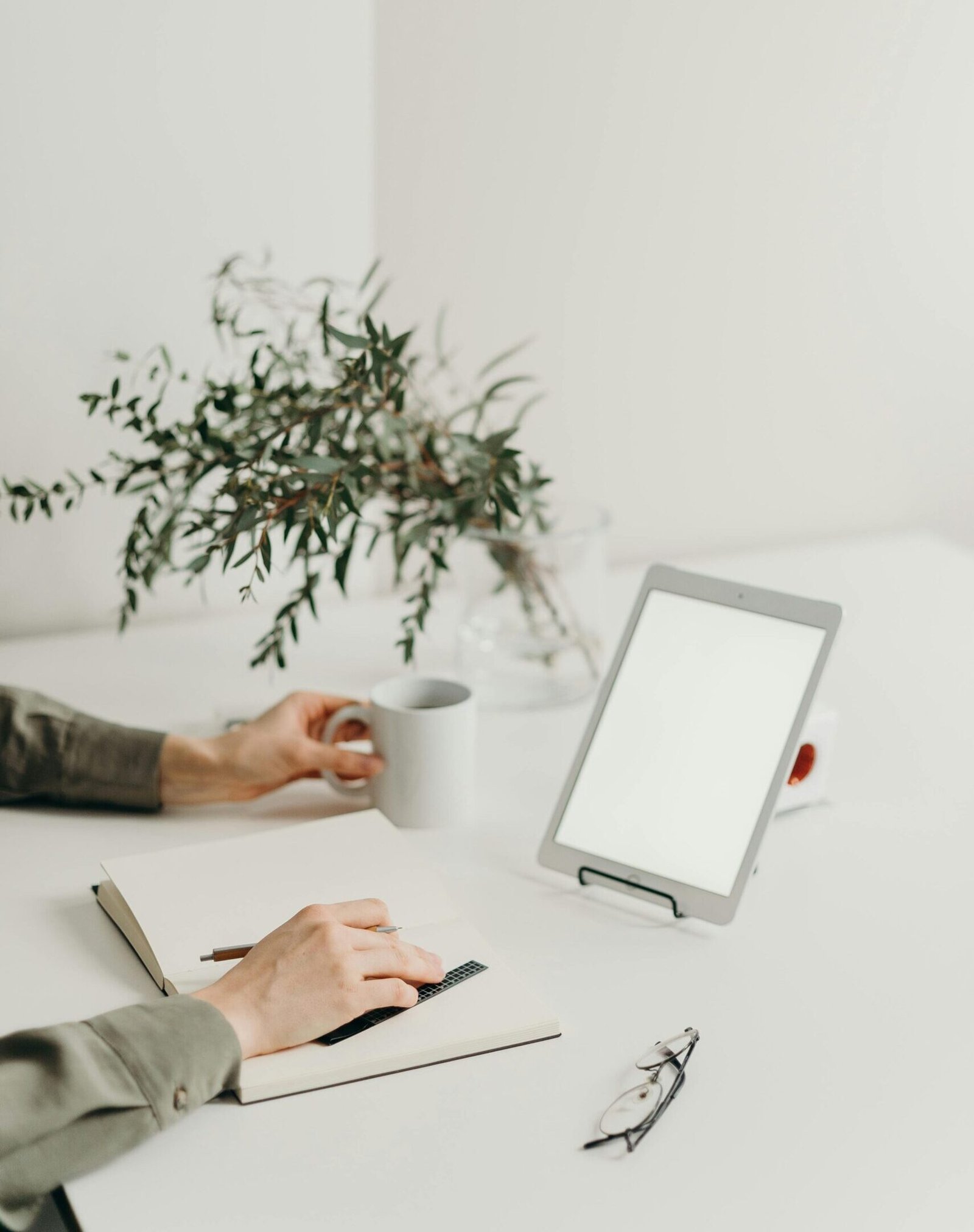 About A minimalist home office setup with a blank screen tablet, coffee mug, and greenery.